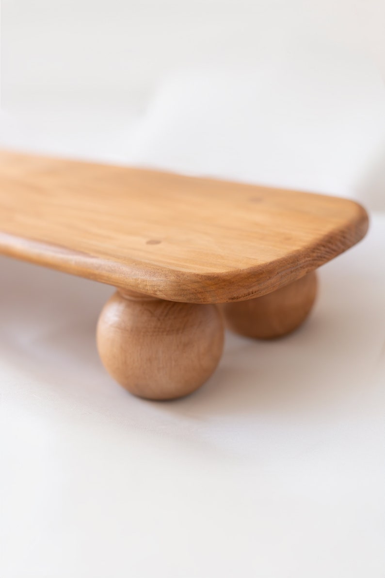 K&ouml;nnte beinhalten: Ein helles Holzserviertablett mit abgerundeten Kanten, das von drei kugelf&ouml;rmigen Holzf&uuml;&szlig;en getragen wird. Das Tablett hat eine nat&uuml;rliche Holzmaserung und eine glatte Oberfl&auml;che, ideal f&uuml;r die Pr&auml;sentation von Speisen oder Dekorationen.