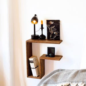 May include: A two-tiered wooden wall shelf. The top shelf displays a black desk lamp, a black candlestick with a lit orange candle, and a framed artwork. The lower shelf holds books and a small black device. A grey and white patterned blanket is below.