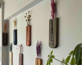 May include: A collection of wall-mounted wooden bud vases, each featuring a vertical slot holding a glass tube with dried flowers. The vases are finished in various wood tones, including light, medium, and dark stains, and hang by thin cords.