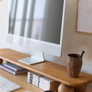 K&ouml;nnte beinhalten: Ein moderner Schreibtisch mit einem wei&szlig;en Computermonitor auf einem Holzst&auml;nder. Ein Holz-Stifthalter, B&uuml;cher, eine Kaffeetasse und eine Computermaus befinden sich ebenfalls auf dem Schreibtisch. Der Schreibtisch ist aus hellem Holz.