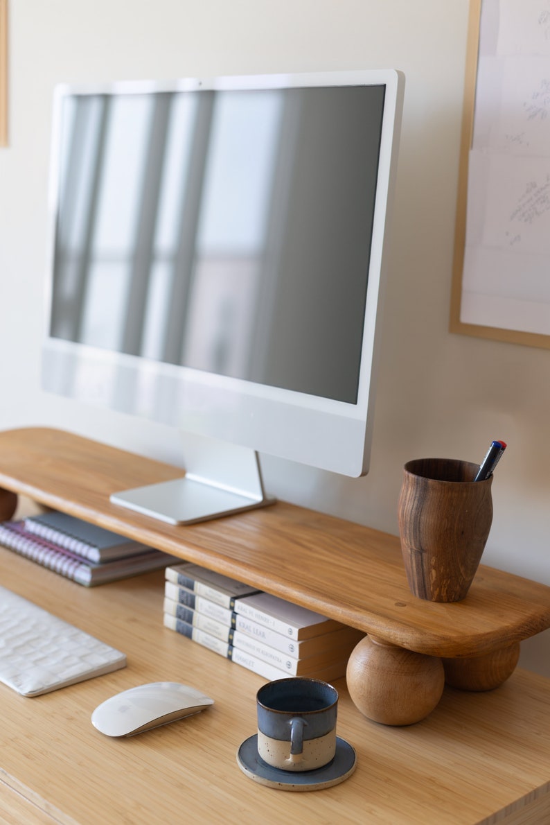 K&ouml;nnte beinhalten: Ein moderner Schreibtisch mit einem wei&szlig;en Computermonitor auf einem Holzst&auml;nder. Ein Holz-Stifthalter, B&uuml;cher, eine Kaffeetasse und eine Computermaus befinden sich ebenfalls auf dem Schreibtisch. Der Schreibtisch ist aus hellem Holz.