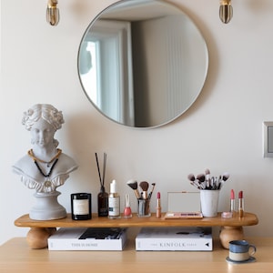 K&ouml;nnte beinhalten: Ein gut beleuchteter Schminktisch mit rundem Spiegel, Holzregal und B&uuml;ste. Das Regal zeigt Make-up-Pinsel, Lippenstifte und Hautpflegeprodukte. Ein Stapel wei&szlig;er Handt&uuml;cher befindet sich auf einem Regal &uuml;ber dem Spiegel.