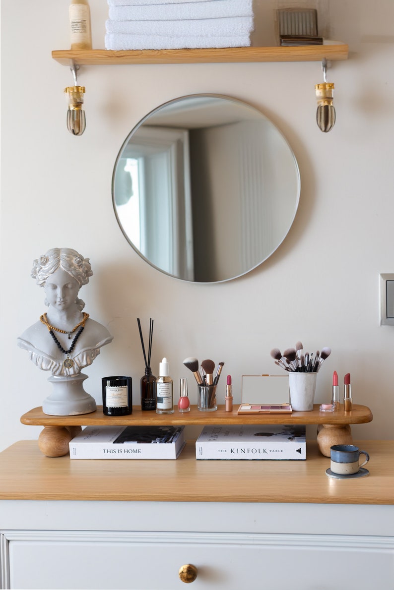 K&ouml;nnte beinhalten: Ein gut beleuchteter Schminktisch mit rundem Spiegel, Holzregal und B&uuml;ste. Das Regal zeigt Make-up-Pinsel, Lippenstifte und Hautpflegeprodukte. Ein Stapel wei&szlig;er Handt&uuml;cher befindet sich auf einem Regal &uuml;ber dem Spiegel.