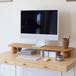 May include: A light wood desk with a computer monitor, wooden monitor stand, and accessories. The desk has three drawers with round knobs. A white keyboard and mouse are on the desk. A small cup and saucer are also on the desk.