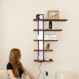 Pode incluir: Uma prateleira de parede de madeira com cinco prateleiras, uma tira de luz e uma variedade de itens nas prateleiras, incluindo livros, um vaso, uma planta e uma moldura de fotos. A prateleira está montada em uma parede branca.