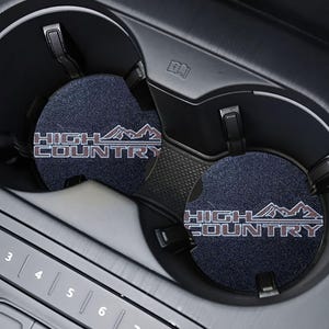 May include: Two black cup coasters with the text "HIGH COUNTRY" in brown lettering and a mountain range graphic. The coasters are in a car's cup holder.