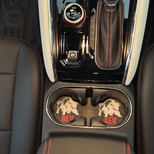 Puede incluir: Dos posavasos negros para coche con un gráfico de toro gris y las letras "AEV" en rojo. Los posavasos están en la consola central de un coche, cerca de la palanca de cambios y los portavasos.