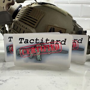 May include: Two white stickers with the text "Tactitard Certified" in black letters. The word "Certified" is stamped in red over a camouflage-patterned rifle.