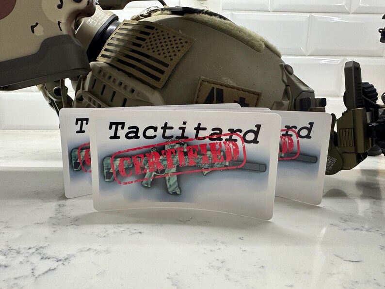 May include: Two white stickers with the text "Tactitard Certified" in black letters. The word "Certified" is stamped in red over a camouflage-patterned rifle.