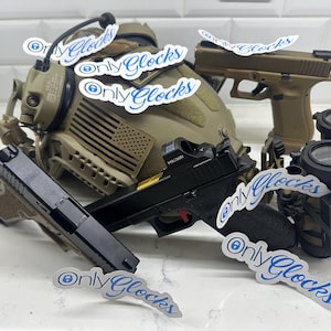 May include: A close-up of a black and tan handgun with a red safety switch and a red dot sight. The gun is lying on a white surface with several white stickers that say "Only Glocks" in blue lettering. There is also a tan helmet with an American flag patch and a pair of black night vision goggles.