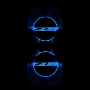 May include: Two illuminated car emblems with the text "ZR2" in a blue glow against a black background. The emblems are circular with a semi-circular design at the top. The text is in a bold, stylized font.