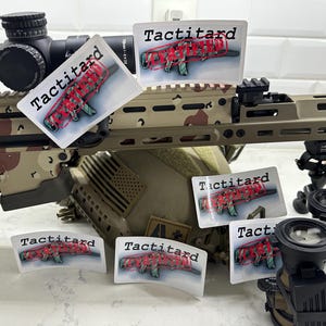 May include: A close-up of a camouflage-patterned rifle with a scope and several stickers that say "Tactitard" with a red line through the word. The stickers are on the rifle, a helmet, and a tripod.