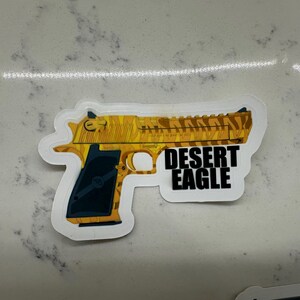 May include: A white sticker with a yellow and black camouflage pattern of a Desert Eagle pistol. The text "DESERT EAGLE" is printed in black letters on the sticker.