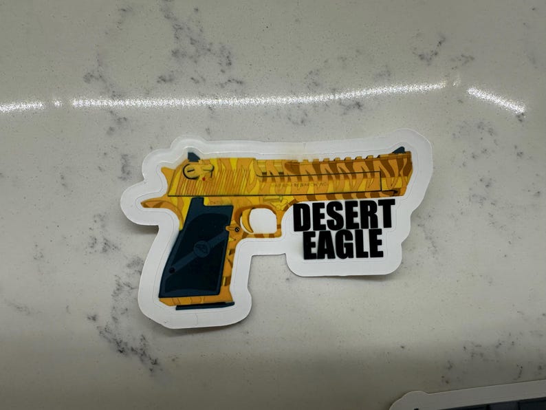 May include: A white sticker with a yellow and black camouflage pattern of a Desert Eagle pistol. The text "DESERT EAGLE" is printed in black letters on the sticker.