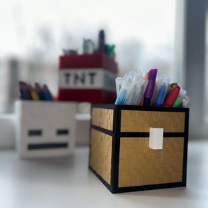 May include: A gold and black pixelated chest-shaped desk organiser filled with pens and markers. A white pixelated skeleton head and a red TNT block organiser are in the background. The chest has a white handle.