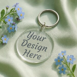 May include: A clear, round keychain with the text "Your Design Here" in gray lettering. The keychain is attached to a silver metal ring. Light blue flowers with yellow centers are scattered around the keychain on a light green satin fabric.