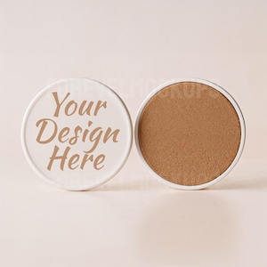 May include: Two round white ceramic coasters with a cork base. One coaster has the text "Your Design Here" in a brown font. The other coaster shows the cork base. The coasters are on a light pink background.