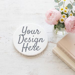 以下が含まれることがあります： 「Your Design Here」の文字がグレーの筆記体で書かれた、白い丸いコースター。白い木の表面に置かれ、ピンクのシャクヤク、デイジー、ユーカリが入った小さな花瓶と、本が積まれたものの隣にあります。