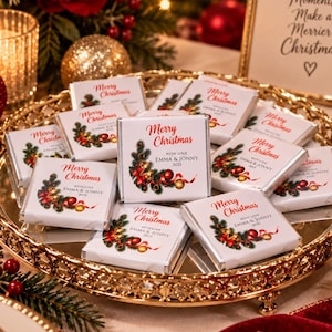 May include: A collection of square chocolate bars wrapped in white paper with a Merry Christmas graphic featuring pine branches, red ornaments, and gold bells. Each bar is printed with the text With Love Emma & Jonny 2025, arranged on a gold tray.