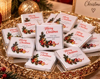 May include: A collection of square chocolate bars wrapped in white paper with a Merry Christmas graphic featuring pine branches, red ornaments, and gold bells. Each bar is printed with the text With Love Emma & Jonny 2025, arranged on a gold tray.