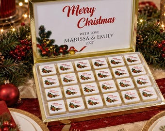 Op de afbeelding: Een goudomrande geschenkdoos met vierentwintig vierkante chocolaatjes, elk versierd met een hulsttakje en de tekst Merry Christmas. Het deksel bevat de tekst Merry Christmas, WITH LOVE, MARISSA & EMILY, en het jaartal 2027.