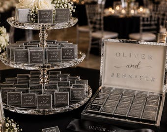 Op de afbeelding: Een kristallen etagère met drie lagen gevuld met vierkante grijze chocolaatjes met het label Oliver and Jennifer, een bijpassende open doos met chocolaatjes, een bordje met Love is Sweet en een zwart lint met de namen.