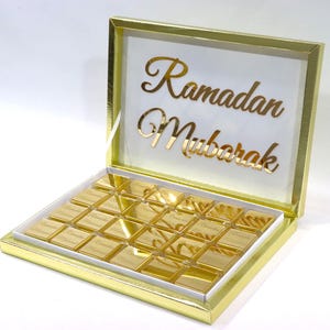 May include: A gold box with a clear lid, filled with square chocolates. The lid displays the words "Ramadan Mubarak" in gold script. The chocolates are individually wrapped in gold foil.
