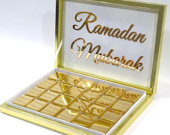 Peut inclure: Une boîte dorée avec un couvercle transparent, contenant des chocolats carrés. Le couvercle porte l'inscription "Ramadan Mubarak" en lettres dorées. Les chocolats sont emballés individuellement dans du papier doré.