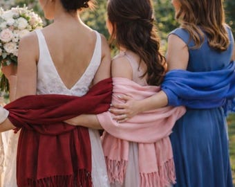 Op de afbeelding: Vier personen staan naast elkaar in mouwloze jurken met gedrapeerde pashmina sjaals in beige, donkerrood, zachtroze en koningsblauw. Elke sjaal is voorzien van een gefranjerde zoom en is om de schouders en armen van de drager gewikkeld.