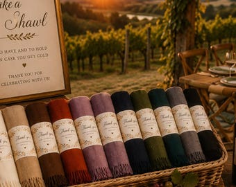 Op de afbeelding: Een rechthoekige rieten mand gevuld met een rij opgerolde sjaals in diverse kleuren, elk omwikkeld met een papieren band met het label Thank You. Een ingelijst bordje vermeldt de tekst Take a Shawl to have and to hold in case you get cold.