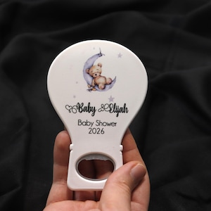 Puede incluir: Abridor de botellas blanco con temática de baby shower. El abridor presenta un osito de peluche en una luna creciente con estrellas. El texto "Baby Elijah Baby Shower 2026" está impreso.