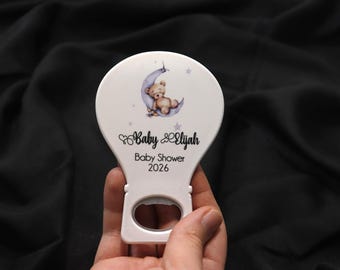 Peut inclure: Décapsuleur blanc sur le thème d'une baby shower. Le décapsuleur représente un ours en peluche sur un croissant de lune avec des étoiles. Le texte "Baby Elijah Baby Shower 2026" est imprimé.