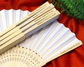 Peut inclure: Plusieurs éventails en papier blanc avec des manches en bambou sont présentés, certains ouverts et d'autres fermés. Les manches sont gravés des noms "Danna & Dean" et de la date "02.24.26". Les éventails sont sur une surface rouge.