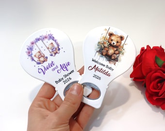 Op de afbeelding: Twee witte flesopeners met baby shower ontwerpen. Eén heeft twee teddyberen op een schommel met de tekst "Violet and Afie Baby Shower 2026". De andere heeft een teddybeer op een schommel met de tekst "Welcome Baby Matilda 2026".