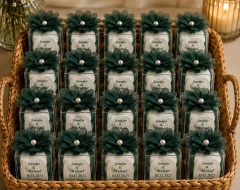 Op de afbeelding: Een verzameling van twintig vierkante doorzichtige doosjes voor bedankjes in een gevlochten mand. Elk doosje bevat witte items en is afgewerkt met een donkergroene stoffen bloem met een witte parel. Labels: Jennifer & Michael 30.11.2025.
