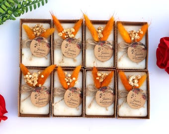 Peut inclure: Huit coffrets cadeaux avec des savons blancs, chacun décoré d'accents orange, de fleurs séchées et d'une étiquette ronde avec l'inscription "Jessica's Bridal Shower". Les boîtes sont disposées sur une surface blanche.