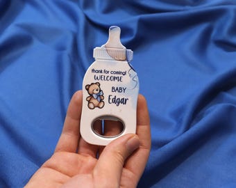 Peut inclure: Un ouvre-bouteille blanc en forme de biberon, avec un motif bleu et blanc. Il présente un dessin d'ours en peluche et le texte "thank for coming! WELCOME BABY Edgar."