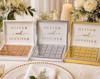 Op de afbeelding: Drie vierkante geschenkdozen met open deksels, elk met twintig kleine vierkante chocolaatjes verpakt in metaalfolie. De binnenkant van elk deksel bevat de gedrukte tekst OLIVER and JENNIFER. De dozen zijn afgewerkt in wit, zilver en goud.