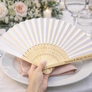 May include: A white and beige folding fan with a wooden handle, resting on a plate with a pink napkin. The fan is personalized with the names "Donna & Dean" and the date "02.24.26".