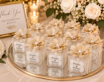 Op de afbeelding: Een verzameling doorzichtige vierkante doosjes voor bedankjes, bekroond met beige stoffen bloemen en witte parels. Elk doosje bevat een wit label met de tekst Jennifer & Michael en de datum 30.11.2025, geplaatst op een ronde spiegel met een gouden rand.