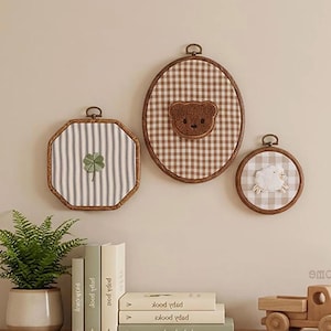 May include: Three wall-mounted embroidery hoops featuring fabric inserts. One octagonal hoop displays a green clover, an oval hoop features a brown teddy bear face, and a small circular hoop shows a white sheep. Each hoop has a wooden frame.