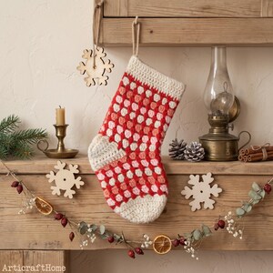 Könnte beinhalten: Ein handgehäkelter Weihnachtsstrumpf mit Granny-Square-Muster in Rot-, Rosa- und Weißtönen. Der Strumpf hat ein cremefarbenes Bündchen sowie eine cremefarbene Ferse und Spitze und ist an einer beigen Schnur an einem Holzregal aufgehängt.