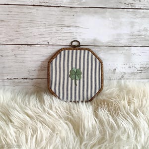May include: An octagonal embroidery hoop frame containing fabric with a blue and white vertical striped pattern. Centered on the fabric is a small, green, four-leaf clover embroidery. The wooden frame has a dark brown finish and a metal hanging loop.