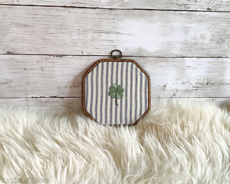 May include: An octagonal embroidery hoop frame containing fabric with a blue and white vertical striped pattern. Centered on the fabric is a small, green, four-leaf clover embroidery. The wooden frame has a dark brown finish and a metal hanging loop.