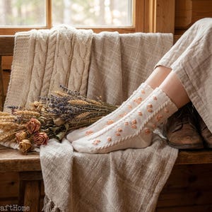 May include: Cream-colored socks with small, peach-colored floral accents. The socks are worn by a person sitting on a wooden bench, with a neutral-toned blanket and dried flowers nearby. The scene is lit by natural light.