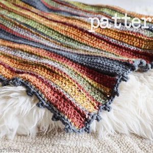 Peut inclure: Une couverture crochetée présentant des rayures verticales dans des tons rouille, vert olive, jaune moutarde et gris anthracite. La couverture a un motif de points côtelés texturés et un bord picot gris anthracite. Le mot pattern est visible.
