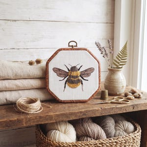 May include: An octagonal wooden embroidery hoop frame containing a white fabric panel with a detailed, textured needlepoint design of a bumblebee with yellow and black stripes and brown wings, featuring a small metal hanging loop at the top.