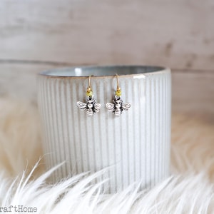 May include: A pair of silver-toned bee earrings with gold-colored hooks and small yellow beads. The earrings are displayed on a light gray ceramic mug with vertical ridges, set against a soft, white, fluffy background.