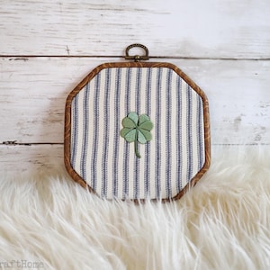 May include: A framed wall hanging with a four-leaf clover design. The frame is octagonal with a brown wood finish. The fabric inside has blue and white vertical stripes. The clover is green with a stem.