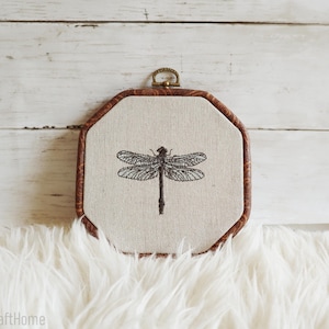 May include: An octagonal embroidery hoop frame with a dark brown wood-grain pattern containing a piece of light beige fabric. Centered on the fabric is a detailed black thread embroidery of a dragonfly with intricate wing patterns.